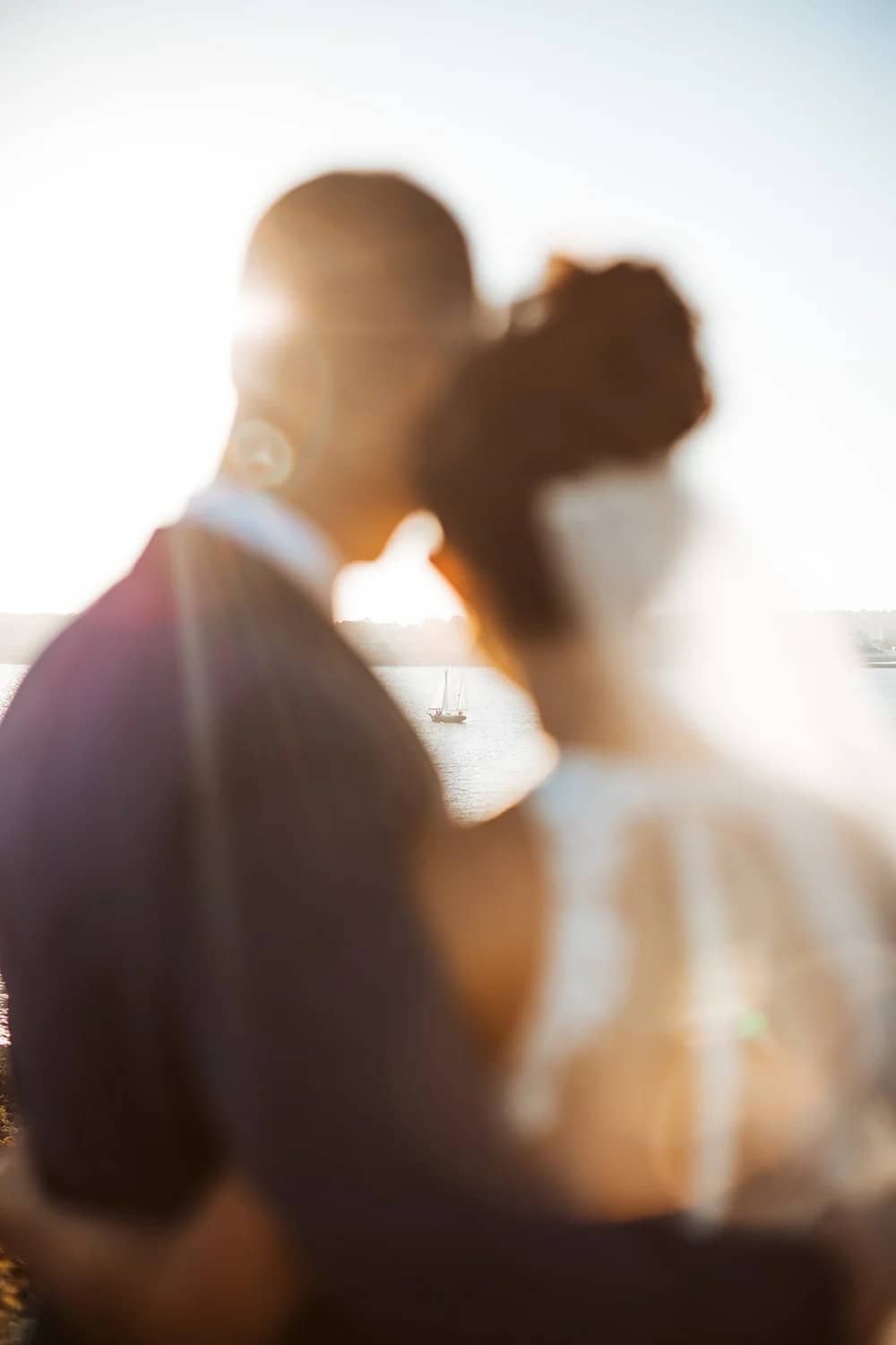 A bride and groom embrace with a sunlit waterfront and sailboat in the background.