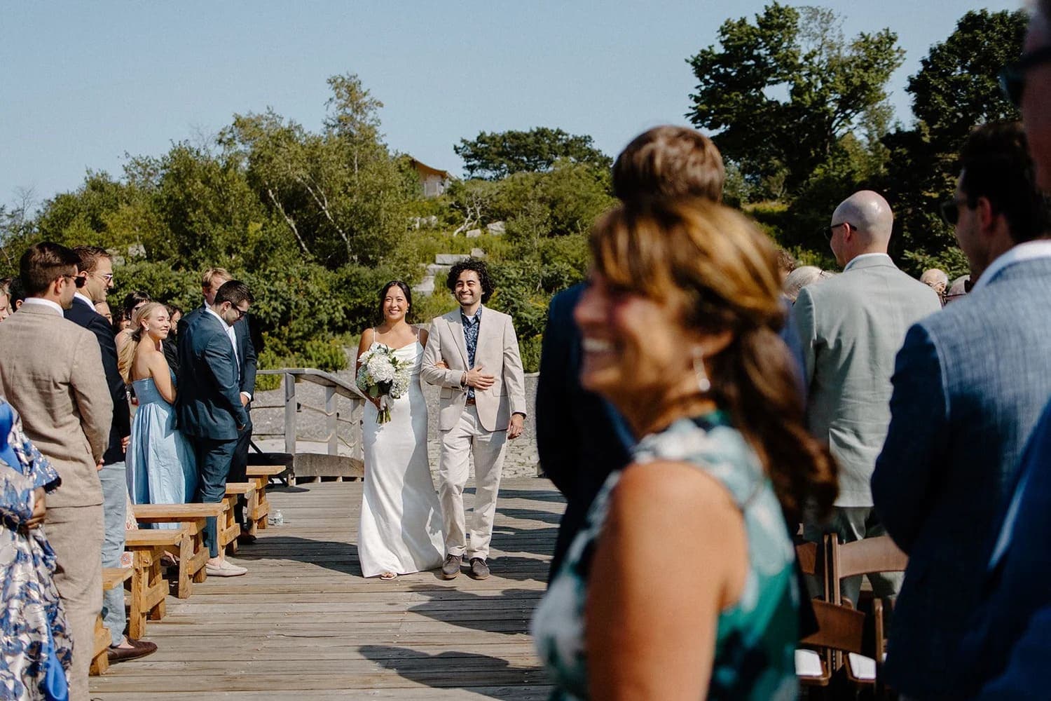 A bride and groom walk down a wooden aisle surrounded by guests at an outdoor wedding.