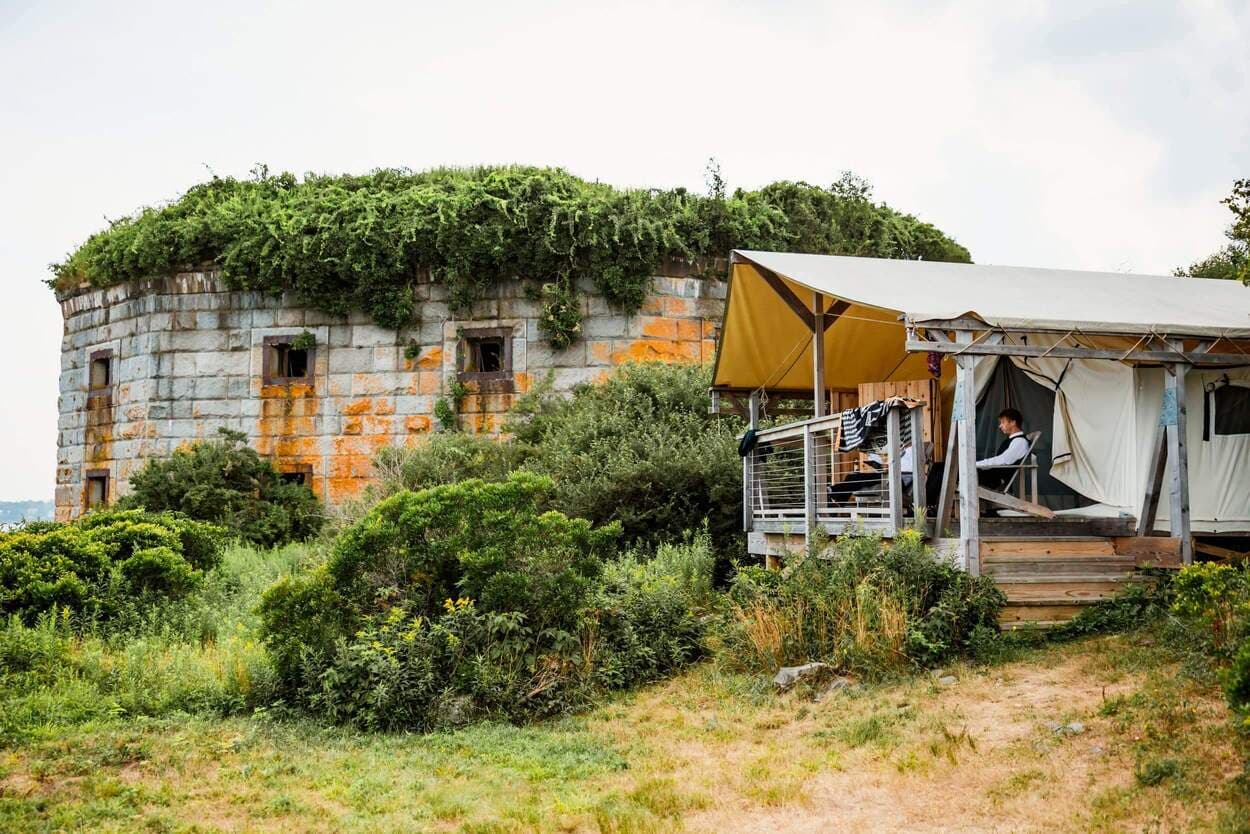A weathered stone structure overgrown with greenery stands beside a tented accommodation in a grassy landscape.