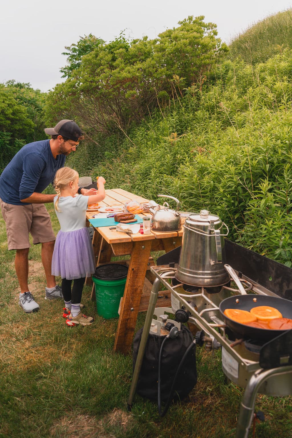 A man and a young girl are preparing food together at an outdoor picnic table.