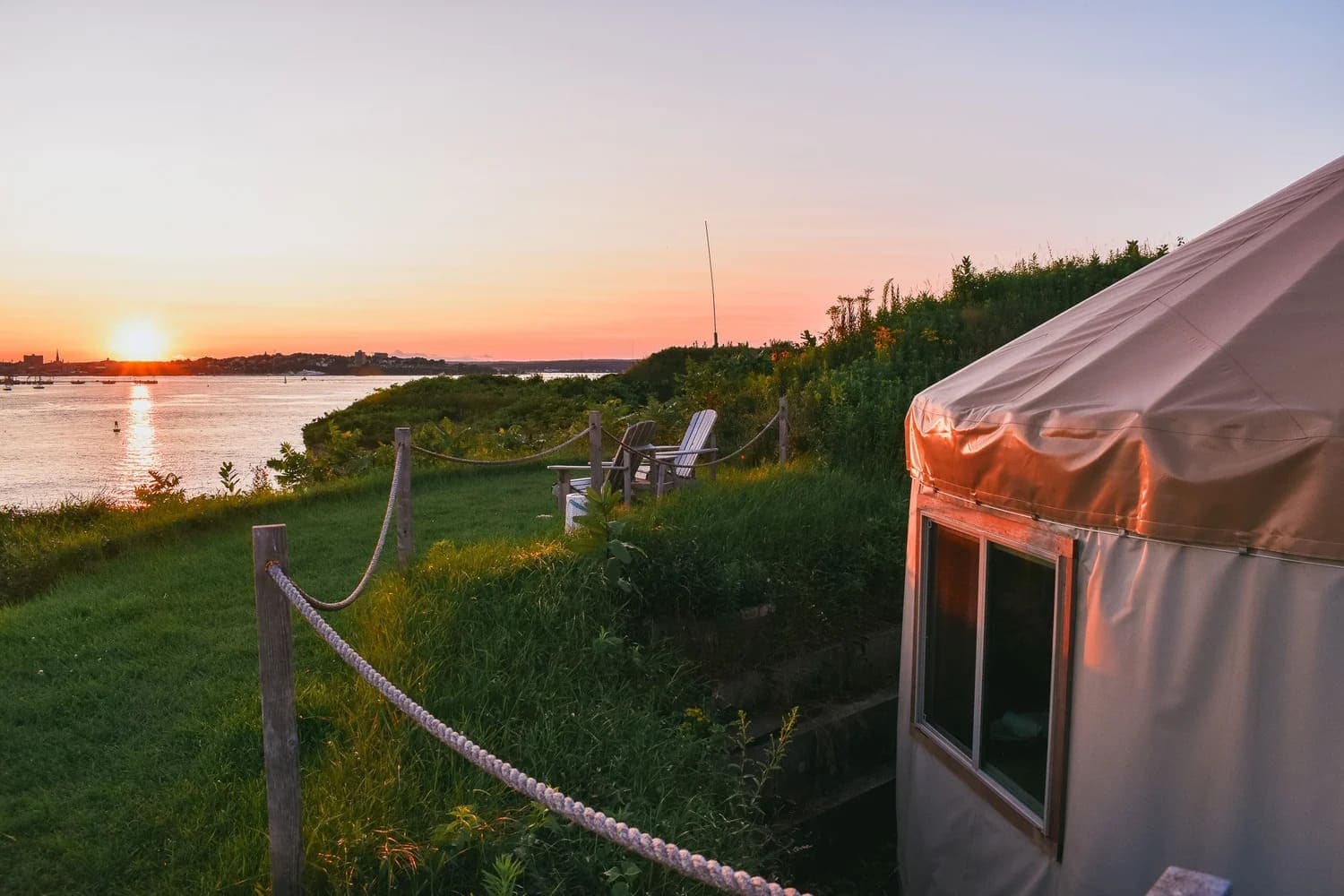 A cozy yurt on a grassy hillside overlooking a serene sunset by the water.