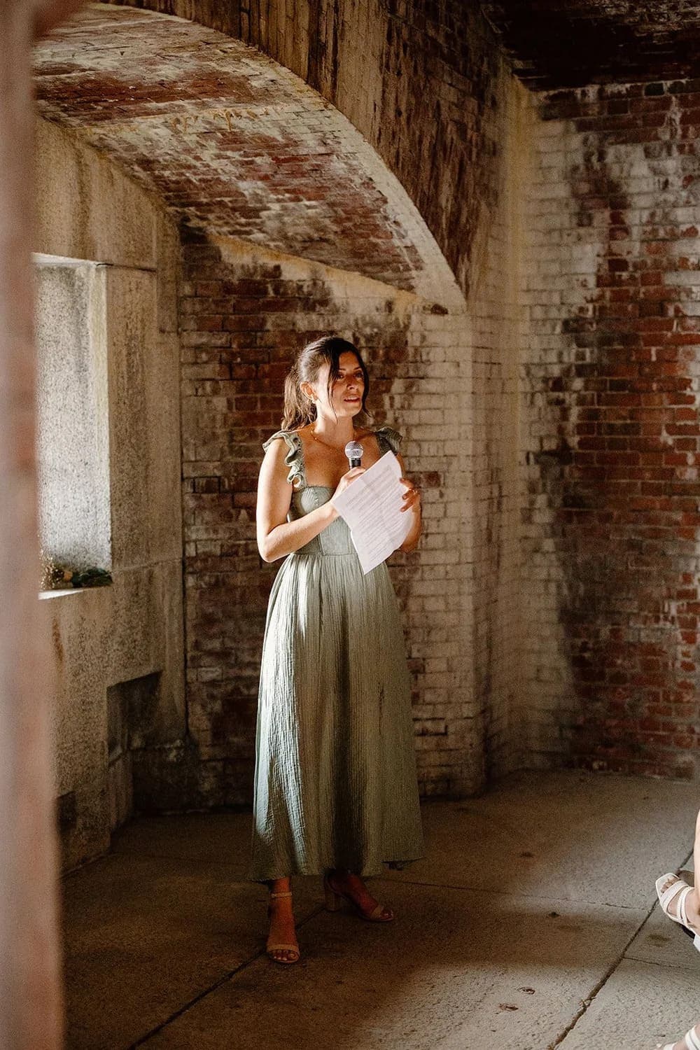 A woman in a green dress speaks into a microphone while holding notes in a brick-walled space.