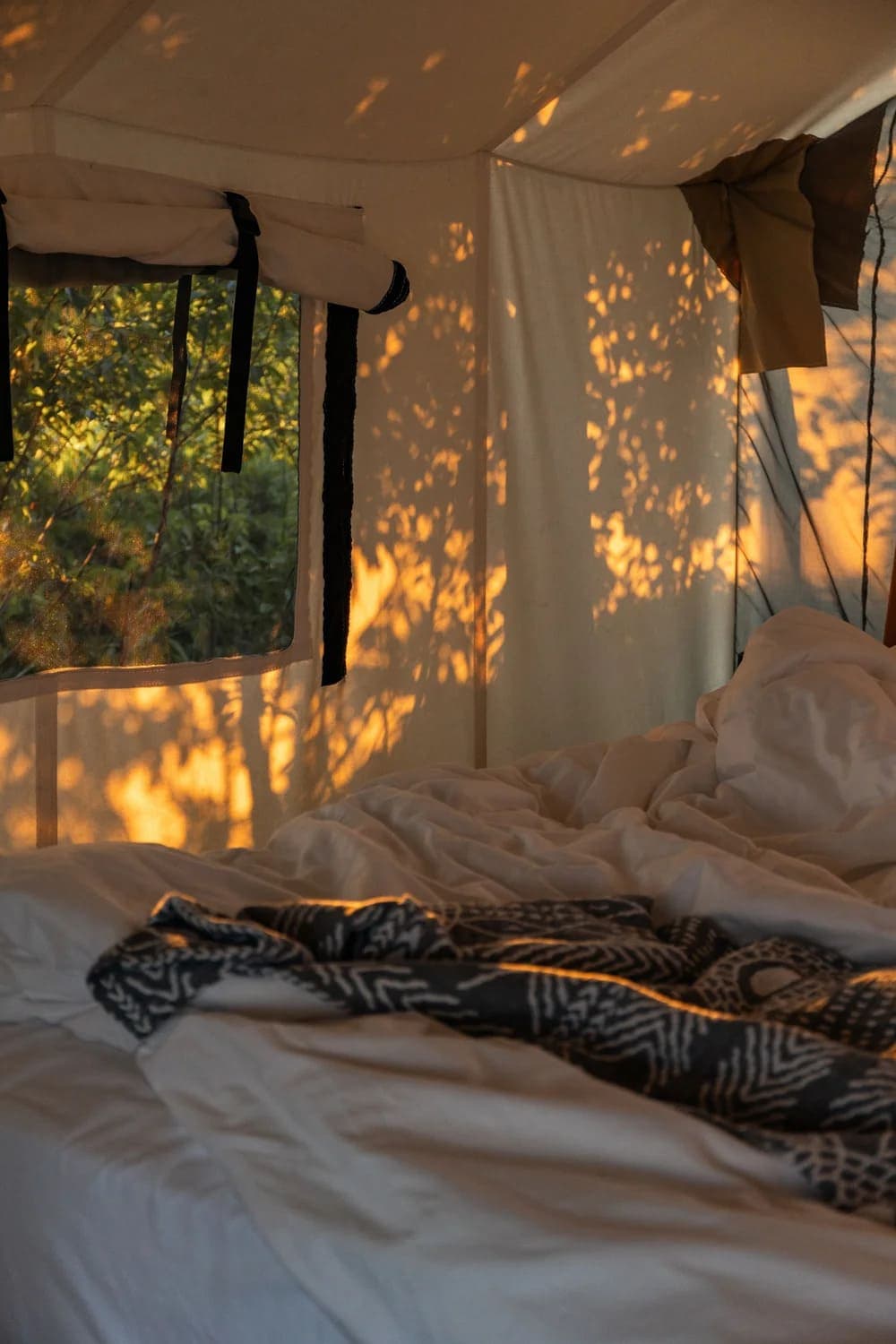 Interior of a tent with sunlight casting shadows on a unmade bed and crumpled blankets.