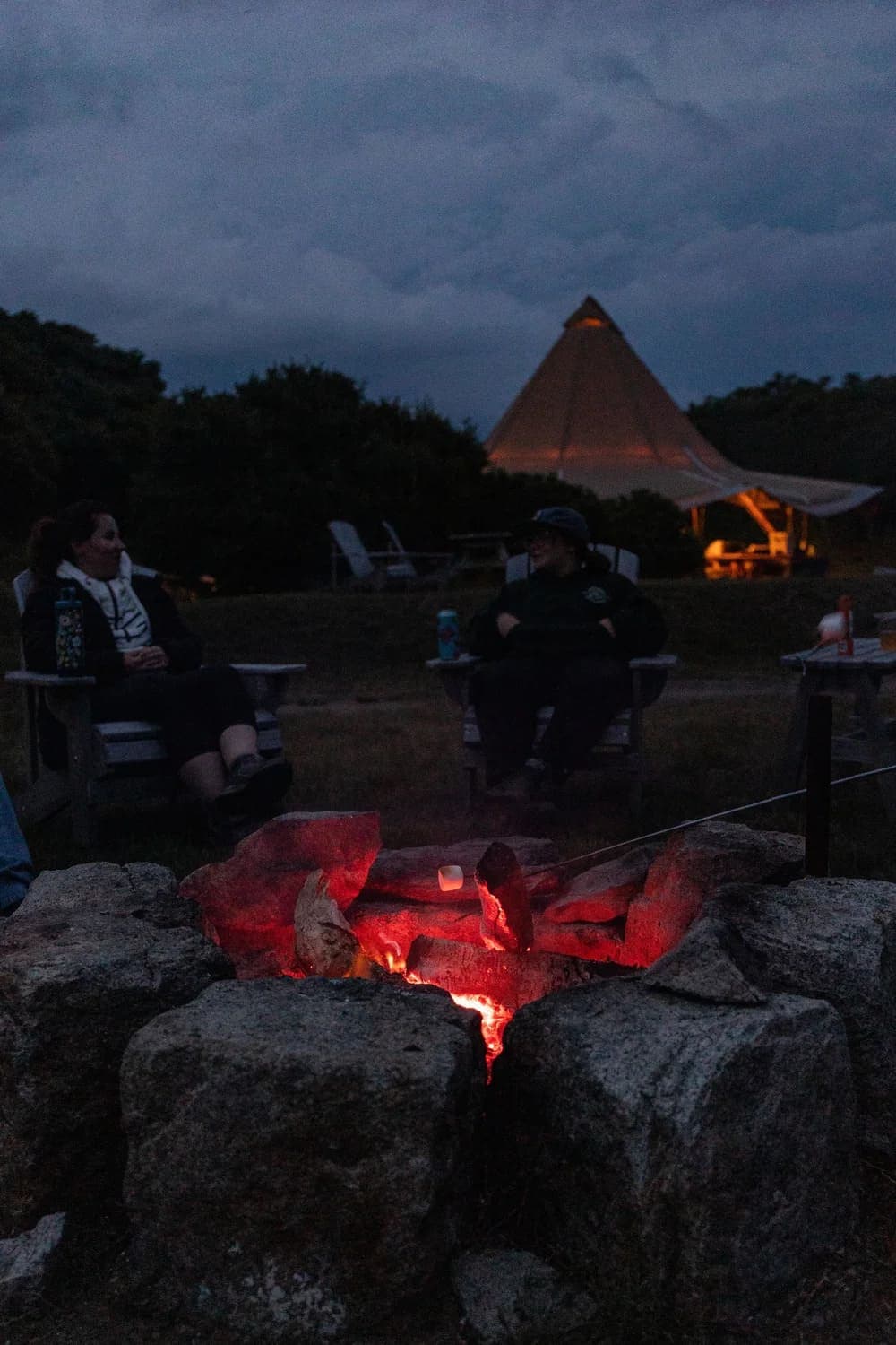 A glowing campfire surrounded by people seated in chairs as night falls.