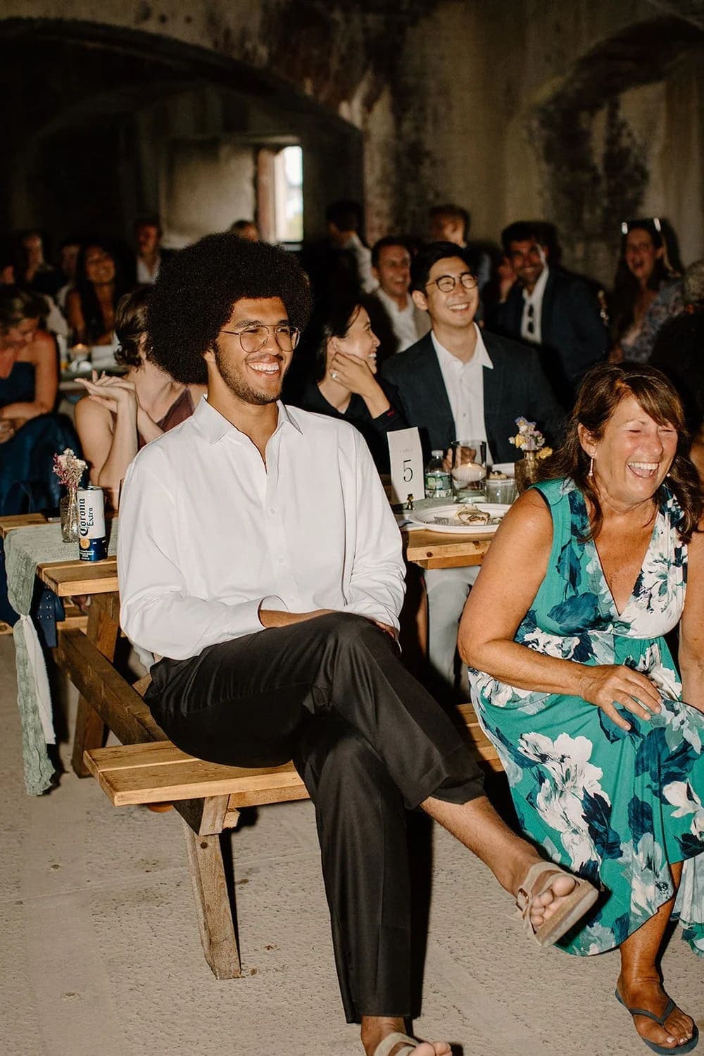 A man with curly hair smiles while seated at a table during a lively gathering, alongside a woman in a floral dress laughing.