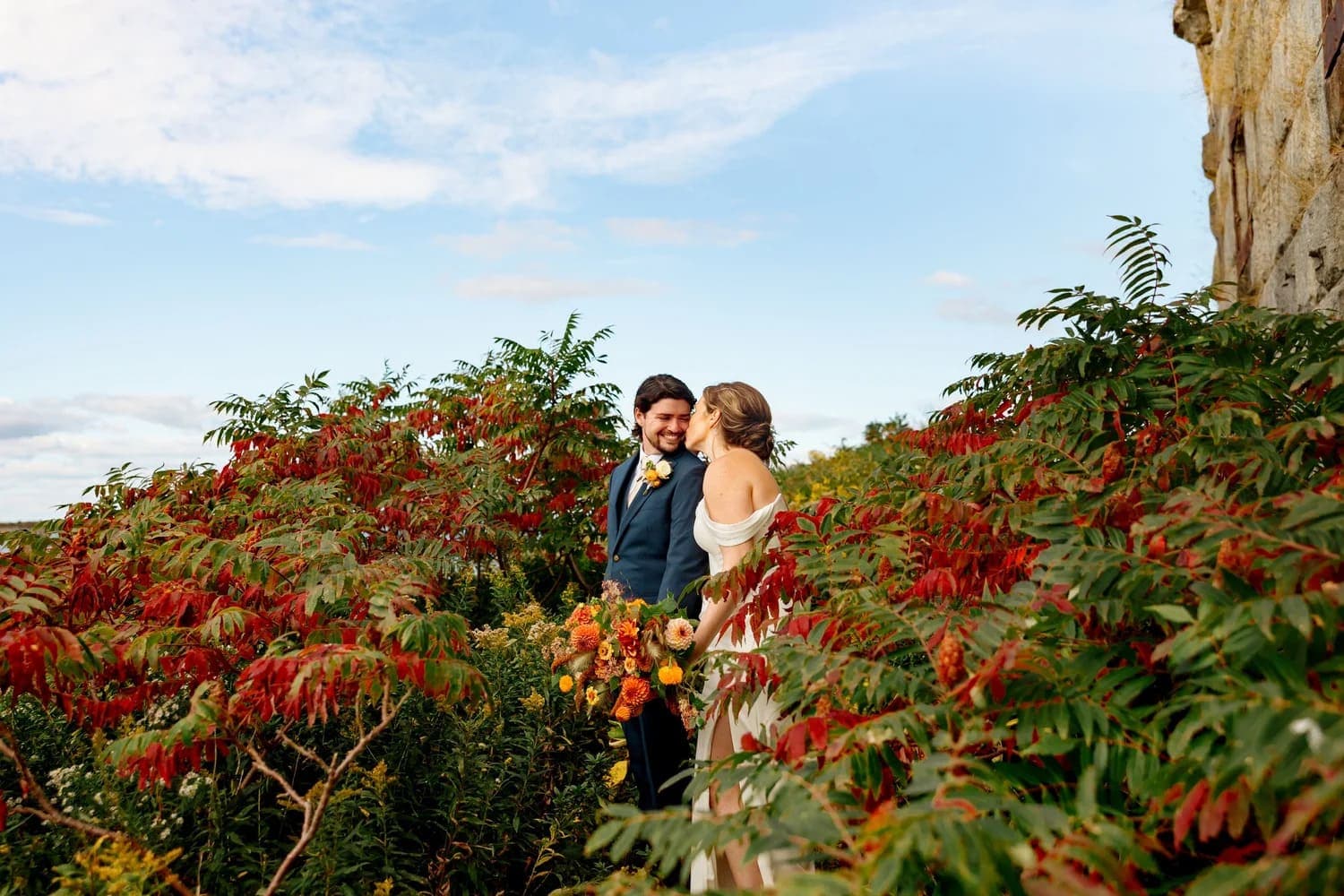 A couple shares a joyful moment surrounded by lush green and red foliage against a picturesque sky.