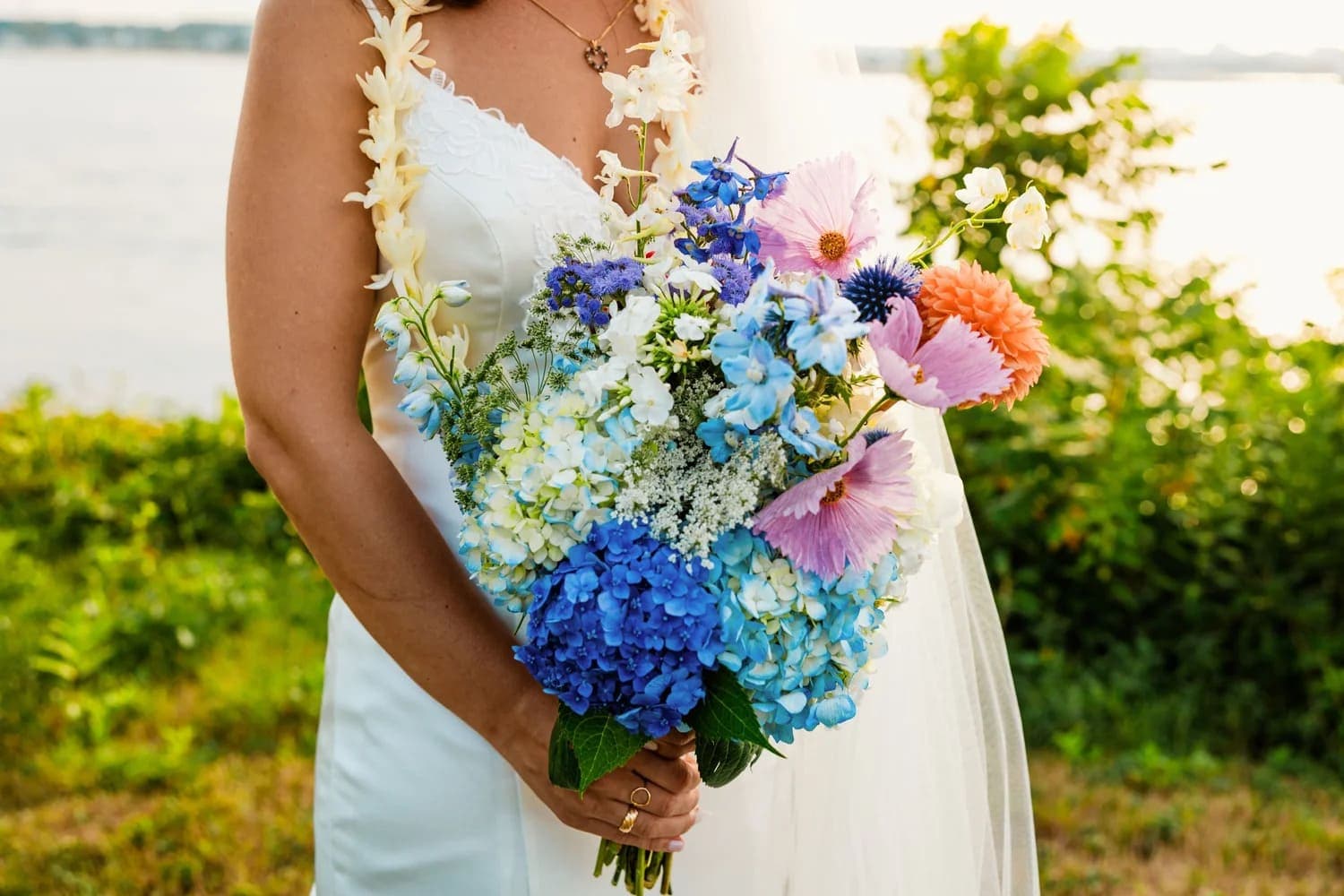 A bride holds a vibrant bouquet of blue and pink flowers against a scenic outdoor background.