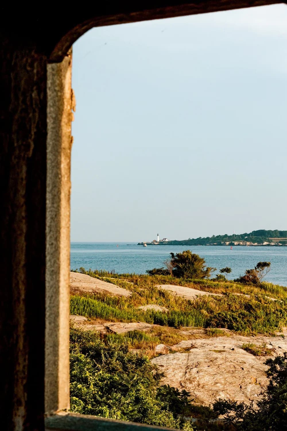 View of a lighthouse through a stone window frame overlooking a calm ocean.
