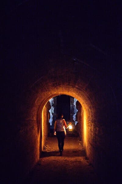 A man walking through underground fort tunnels lit only by the lantern he's holding